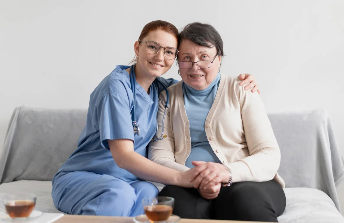 A nurse in blue scrubs sits closely and provides emotional support to an elderly woman on a couch, both holding hands.