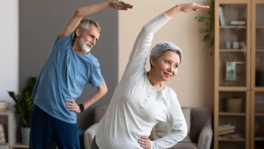 Elderly woman and man stretching arms together at home.