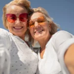 Two older women taking a cheerful selfie together under a clear blue sky.