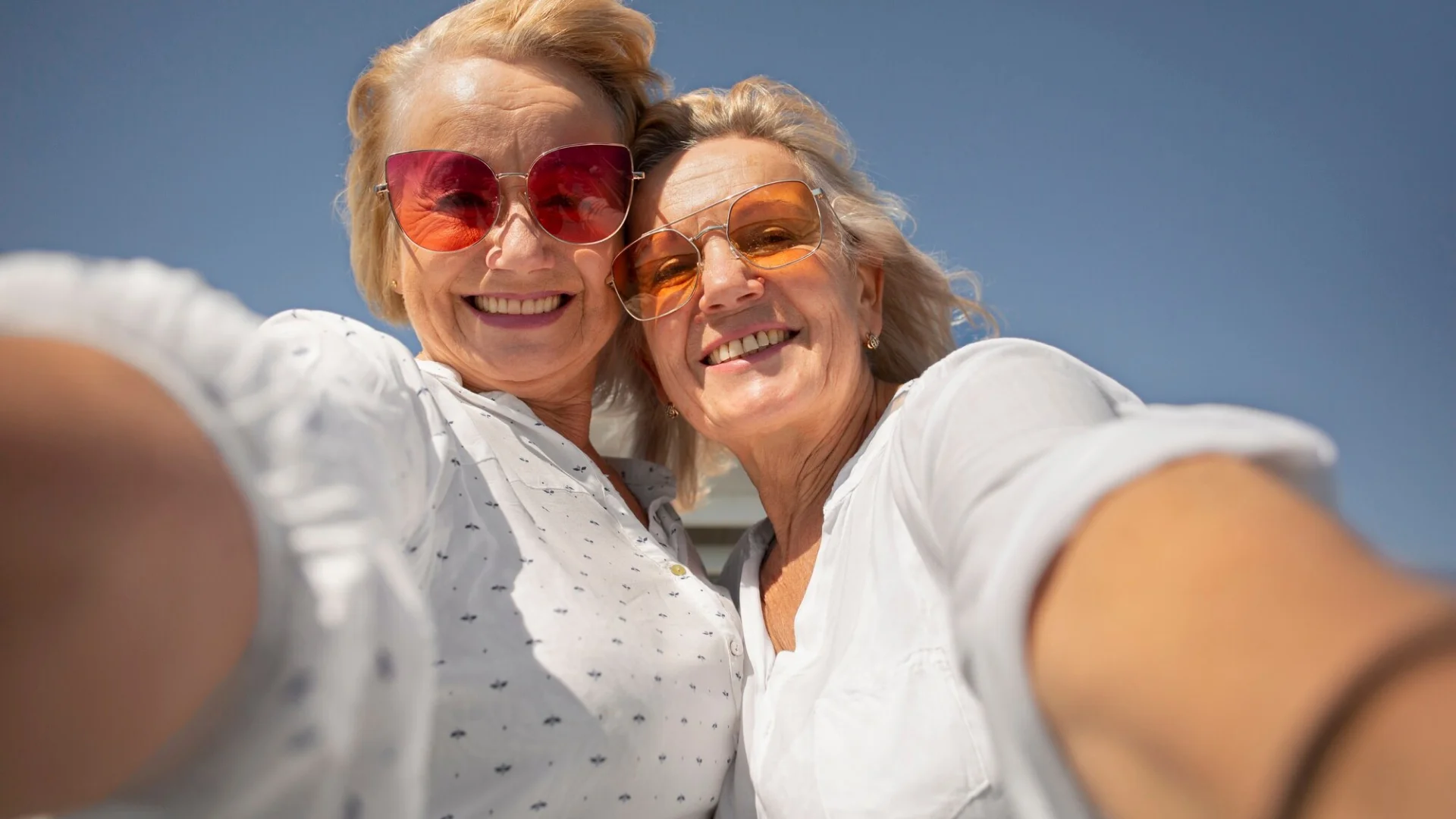 Two older women taking a cheerful selfie together under a clear blue sky.