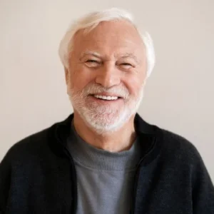 An elderly man with short, white hair is wearing a grey shirt and a black cardigan, photographed against a plain light background.