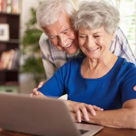 An elderly woman in a blue shirt is sitting at a desk using a laptop, while another older adult stands beside her, gently supporting and looking at the screen.
