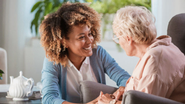 A caregiver offers emotional support and holds hands with an elderly woman, having a comforting conversation in a bright living room.