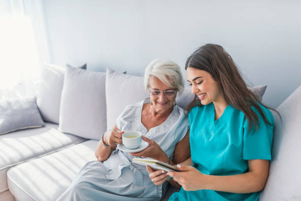 Happy patient is holding caregiver for a hand while spending time together. Elderly woman in nursing home and nurse. Aged elegant woman and tea time at nursing home