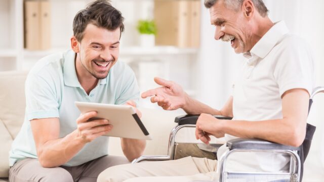 A caregiver sits and discusses something on a tablet with an elderly man in a wheelchair in a bright room.