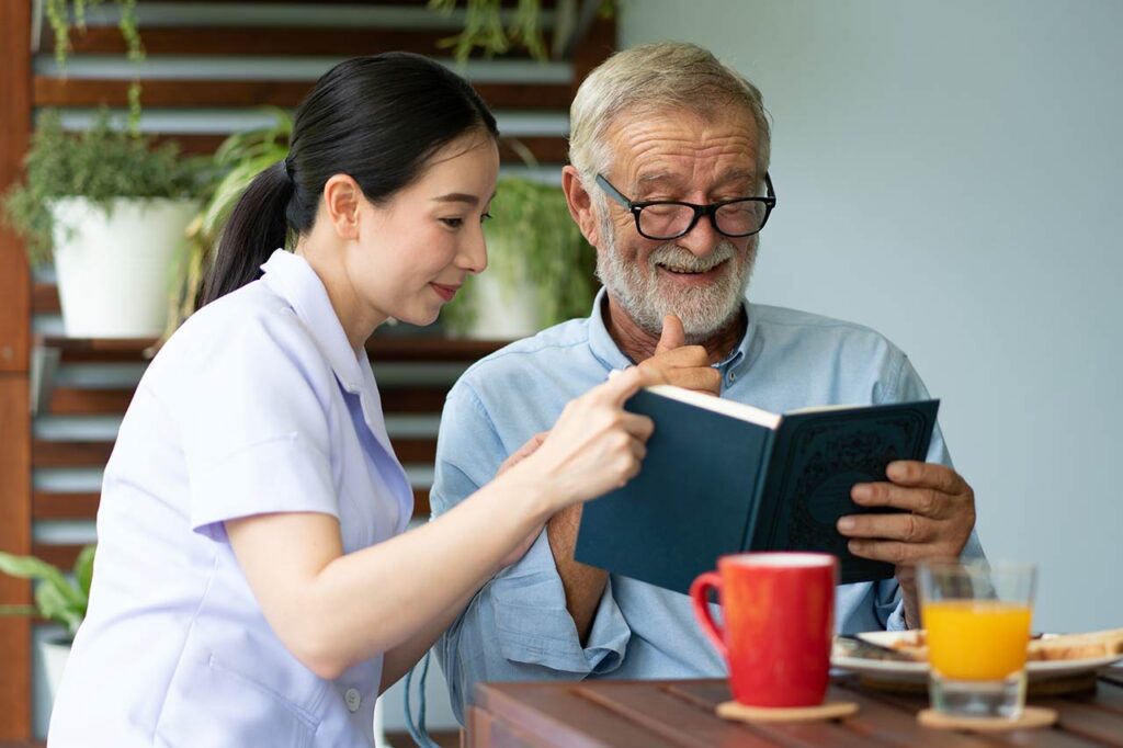 A caregiver discuss a book with an elderly man over a cup of tea in a comfortable indoor setting.