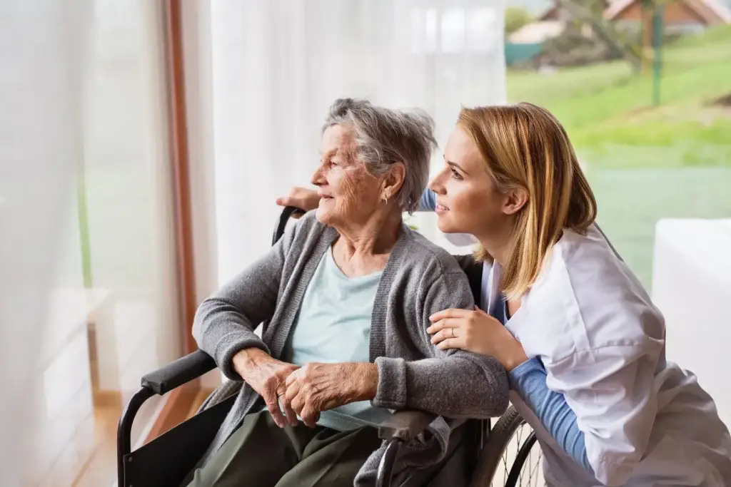 A caregiver in a white coat and an elderly woman in a wheelchair enjoy a peaceful moment together, looking out the window.