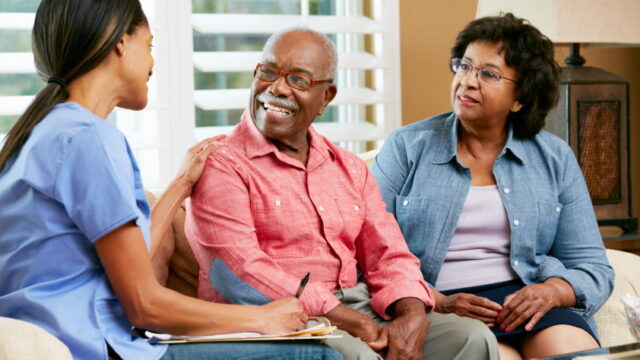 A nurse in blue scrubs talks with an elderly man and woman, both smiling, during a meeting in a comfortable living room.