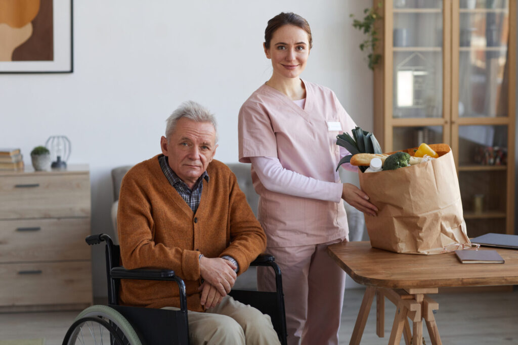 A caregiver in pink scrubs stands beside an elderly man in a wheelchair, holding a grocery bag filled with fresh vegetables.