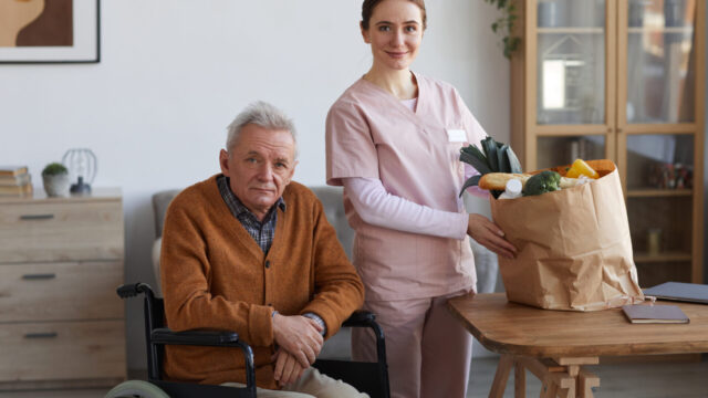 A caregiver in pink scrubs stands beside an elderly man in a wheelchair, holding a grocery bag filled with fresh vegetables.
