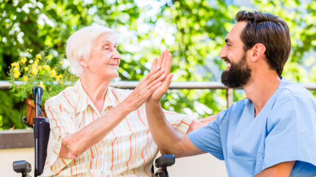 A nurse in blue scrubs exchanges a cheerful high-five with an elderly woman in a striped dress who is sitting in a wheelchair outdoors.
