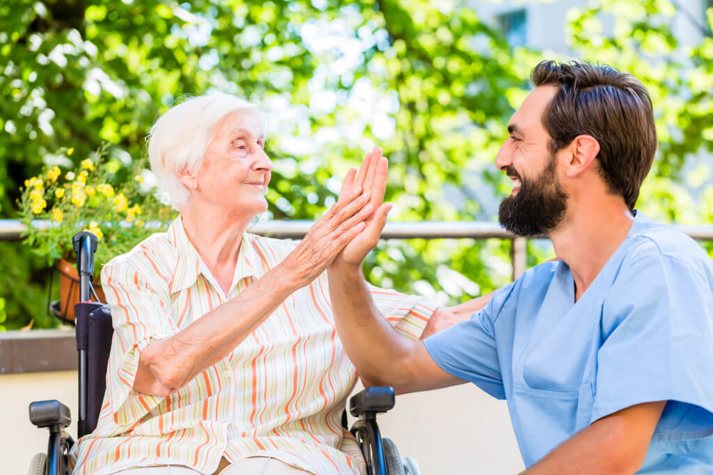 A nurse in blue scrubs exchanges a cheerful high-five with an elderly woman in a striped dress who is sitting in a wheelchair outdoors.