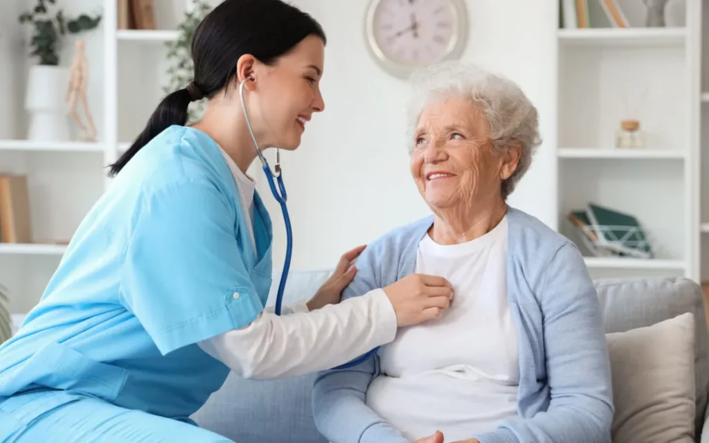 A caregiver in blue scrubs smiles at an elderly woman while adjusting a stethoscope on her chest.