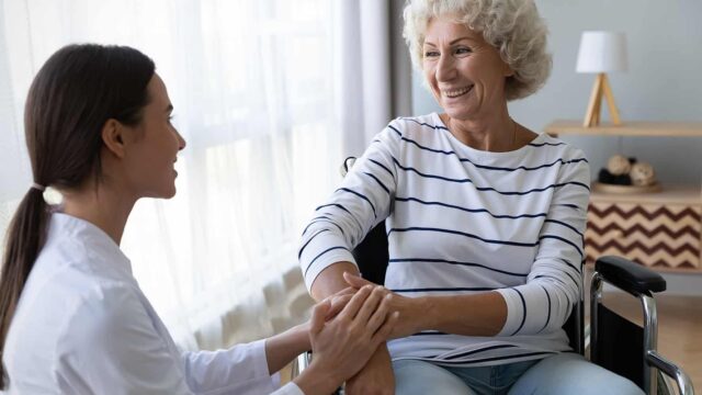 A caregiver in white scrubs holds hands with an elderly woman in a wheelchair, sharing a joyful moment together.