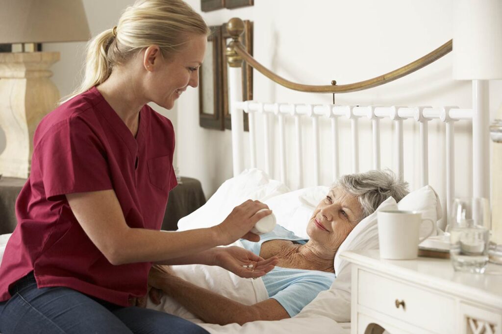 A nurse in a red uniform helps give medication to an elderly woman who is lying in bed in a comfortable room.