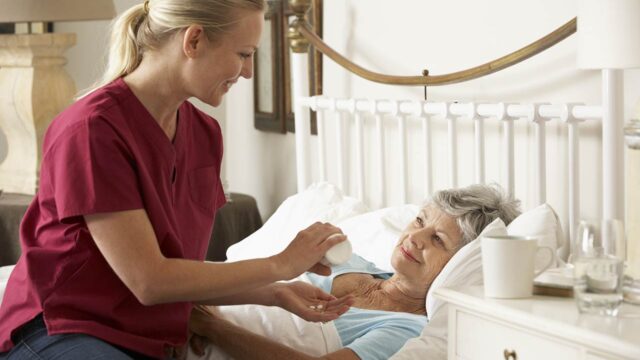 A nurse in a red uniform helps give medication to an elderly woman who is lying in bed in a comfortable room.