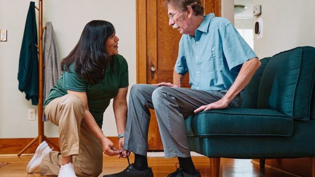 A caregiver helps an elderly man put on his socks while seated on a couch.
