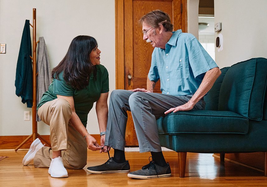 A caregiver helps an elderly man put on his socks while seated on a couch.