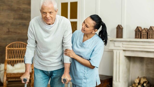 Nurse helping elderly man stand with walker at home.