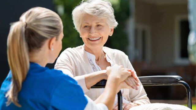 Nurse chatting with elderly woman in wheelchair outdoors.