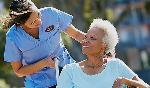 A nurse in blue scrubs pushes an elderly woman in a wheelchair during a sunny day outdoors.
