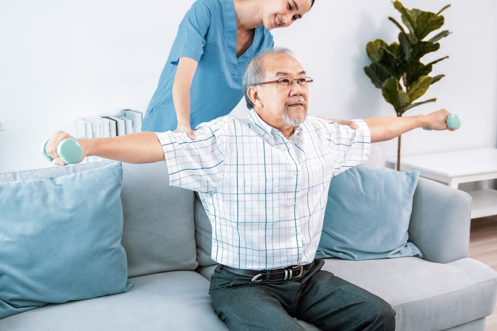 A nurse in blue scrubs assists an elderly man in a checkered shirt as he exercises with small dumbbells on a couch.
