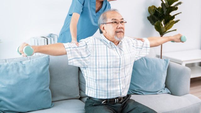 A nurse in blue scrubs assists an elderly man in a checkered shirt as he exercises with small dumbbells on a couch.