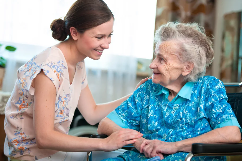 A caregiver smiles at an elderly woman in a wheelchair, sharing a warm and caring moment together.