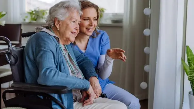 A caregiver in blue scrubs and an elderly woman in a wheelchair share a joyful moment while looking at something together.