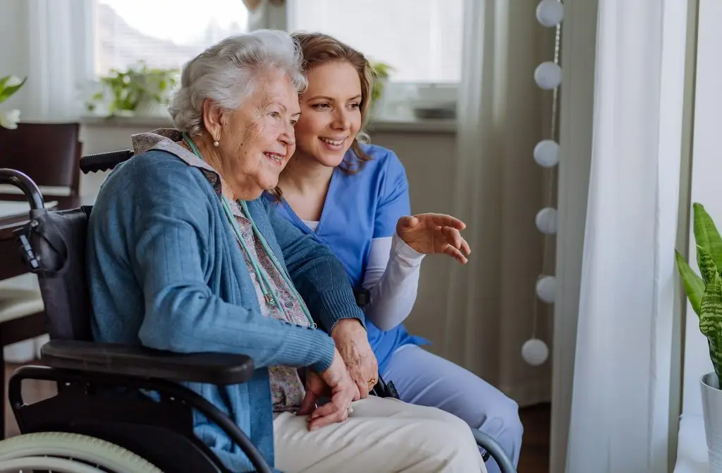 A caregiver in blue scrubs and an elderly woman in a wheelchair share a joyful moment while looking at something together.