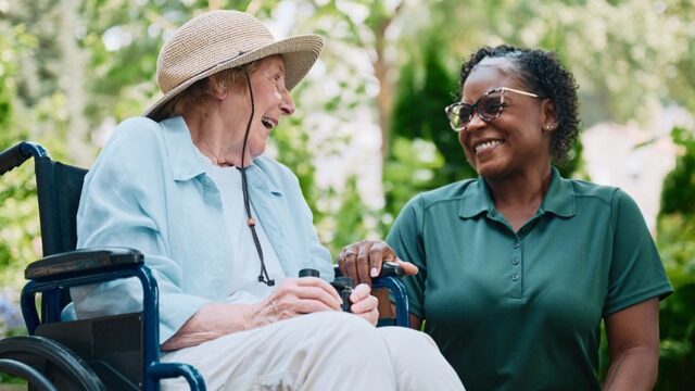 An elderly woman in a wheelchair and a caregiver share a joyful moment outdoors.