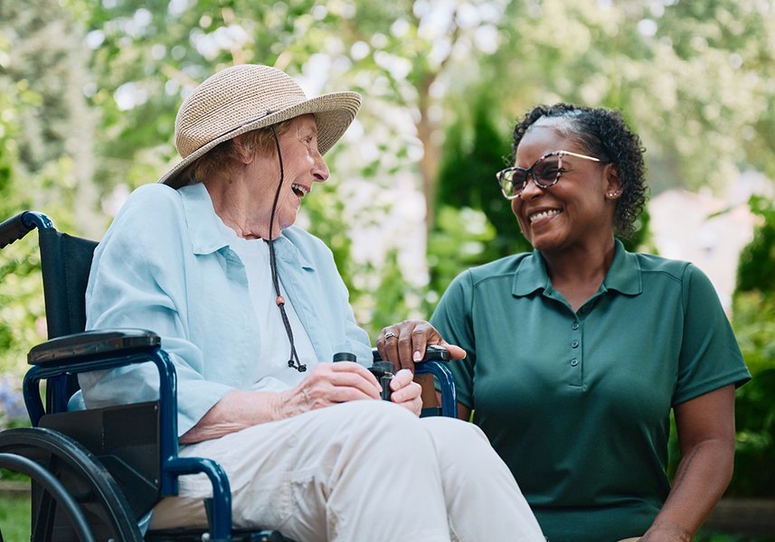 An elderly woman in a wheelchair and a caregiver share a joyful moment outdoors.