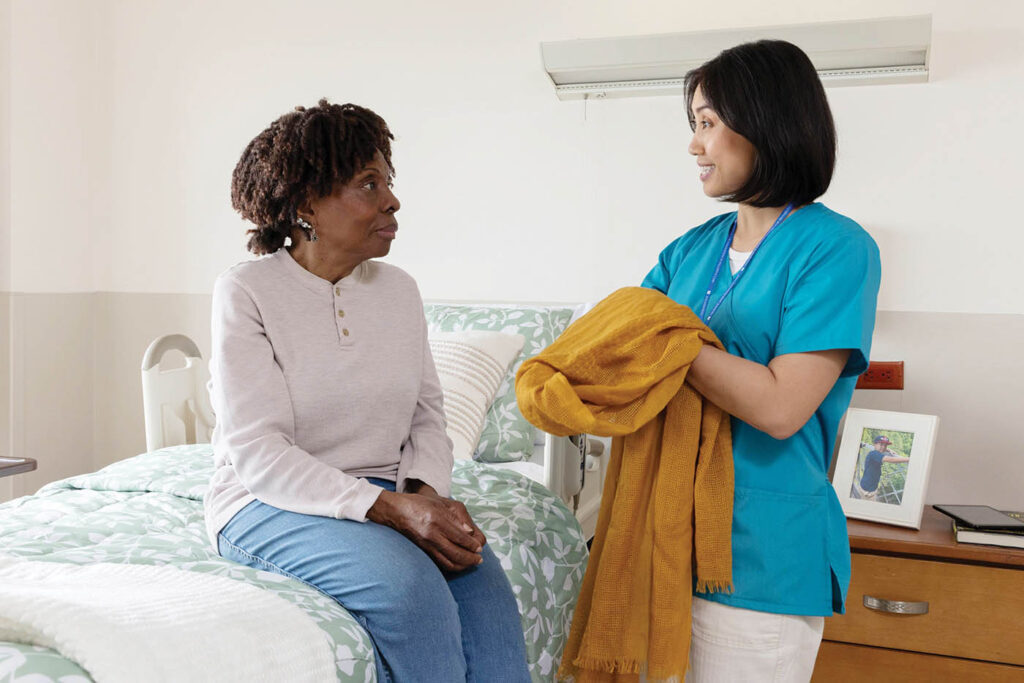 Female caregiver presenting a coat to a seated woman indoors.