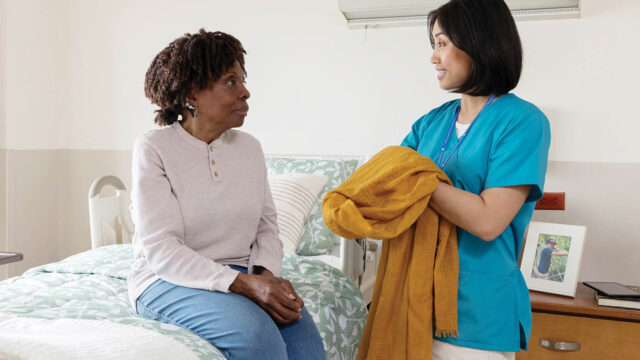 Female caregiver presenting a coat to a seated woman indoors.
