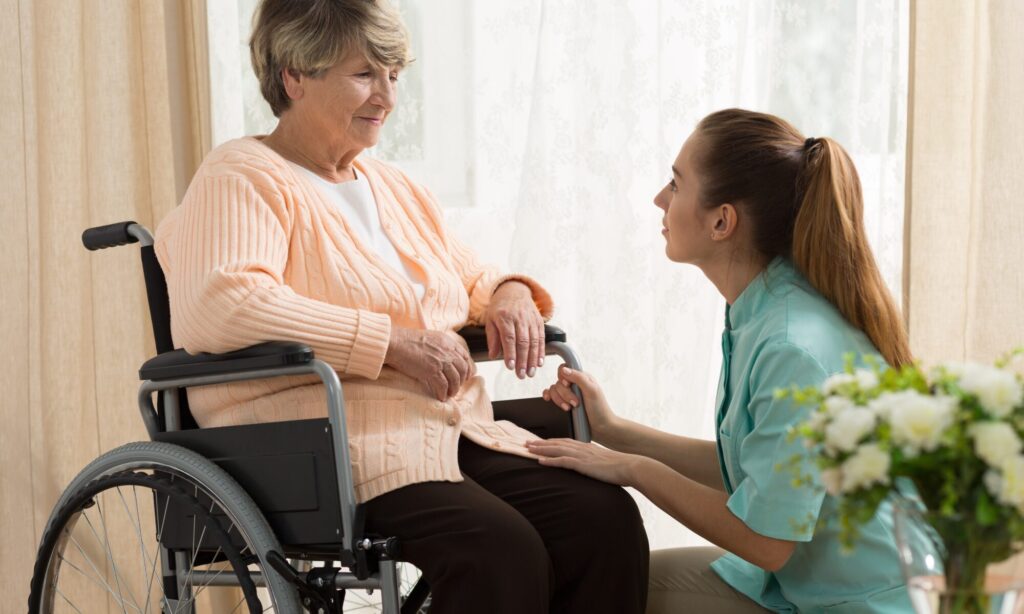 A caregiver helps an elderly woman in a wheelchair with a warm, supportive gesture.