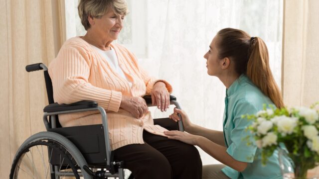 A caregiver helps an elderly woman in a wheelchair with a warm, supportive gesture.