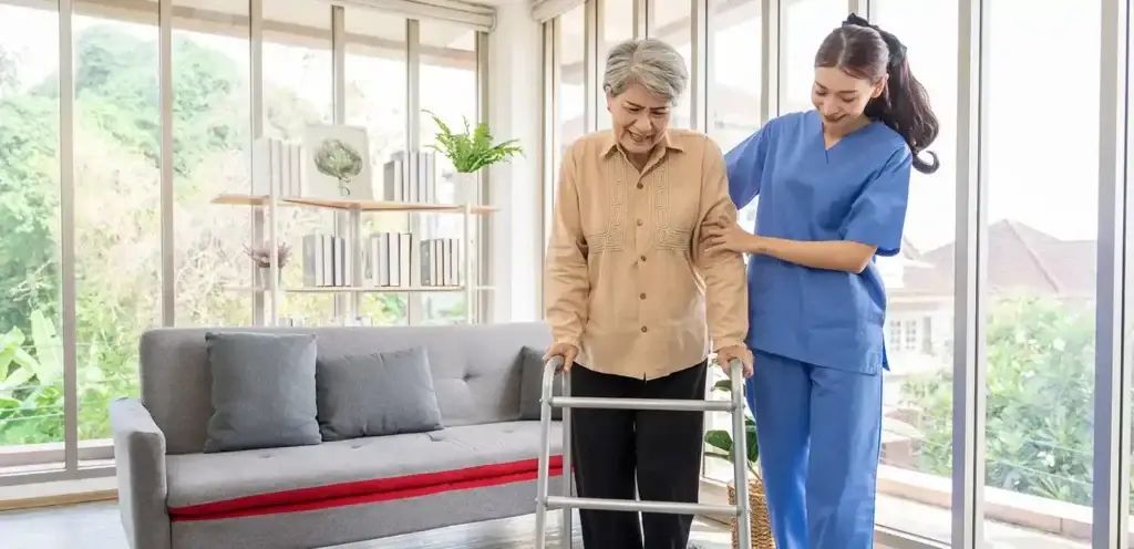 An elderly woman using a walker for support while a caregiver in blue scrubs assists her. The setting is a bright, modern living room with large windows and plants.