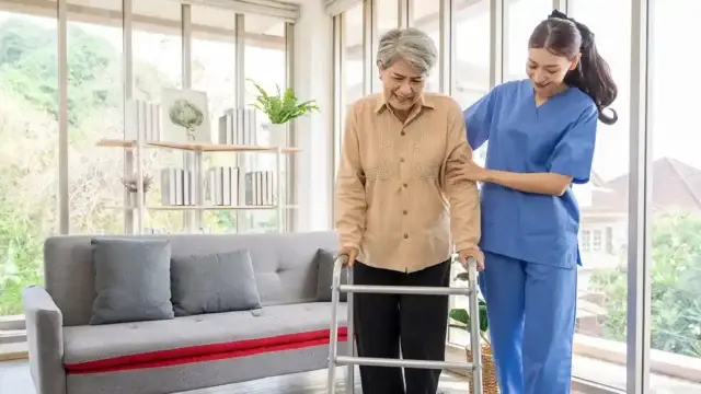 An elderly woman using a walker for support while a caregiver in blue scrubs assists her. The setting is a bright, modern living room with large windows and plants.