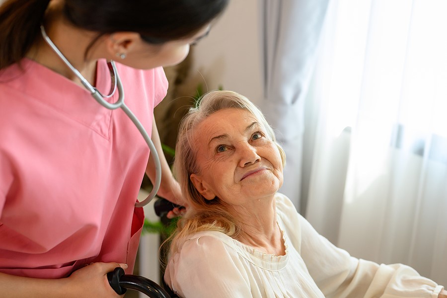 A caregiver in pink scrubs adjusts the stethoscope as she smiles at an elderly woman in a wheelchair.