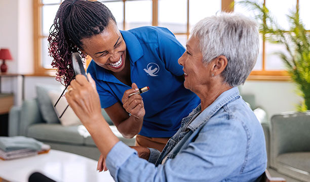 A caregiver in a blue uniform shares a joyful moment with an elderly woman, both smiling.