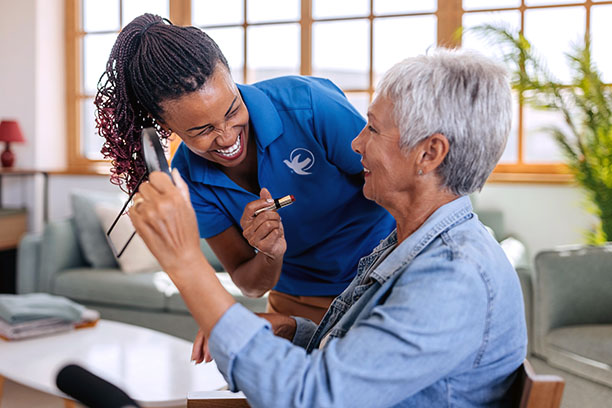 A caregiver in a blue uniform shares a joyful moment with an elderly woman, both smiling.