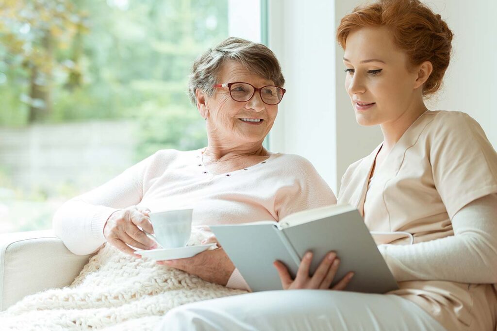 A caregiver in beige scrubs reads a book to an elderly woman holding a cup of tea, both smiling in a cozy setting.