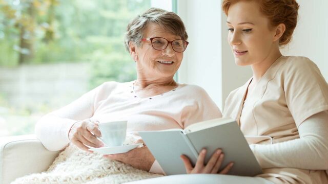 A caregiver in beige scrubs reads a book to an elderly woman holding a cup of tea, both smiling in a cozy setting.