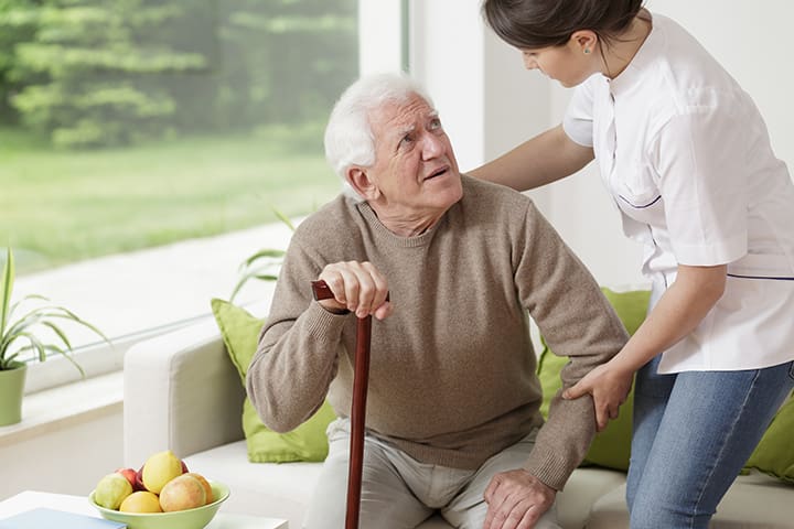 A caregiver assists an elderly man with a cane, offering support as he looks up at her.