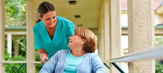A caregiver in green scrubs helps an elderly woman in a wheelchair, both smiling as they share a moment outdoors.