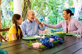 Three people sit at a table outdoors, smiling and sharing a meal together on a bright day.