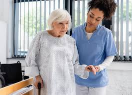 A nurse in blue scrubs gently guides an elderly woman walking with a cane in a bright room.