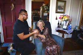 A man kneels and holds the hands of an elderly woman sitting in an armchair in a cozy living room.