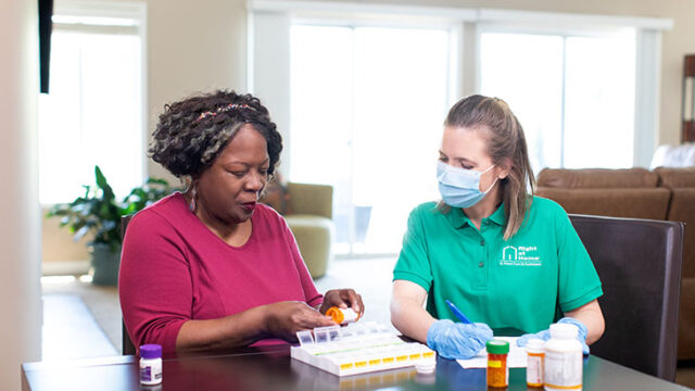 A caregiver in a green shirt assists an elderly woman with organizing her medications into a pill box.