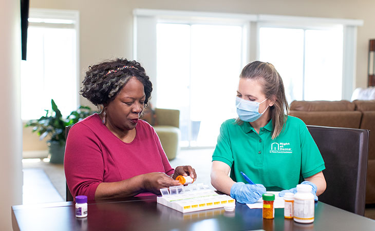 A caregiver in a green shirt assists an elderly woman with organizing her medications into a pill box.